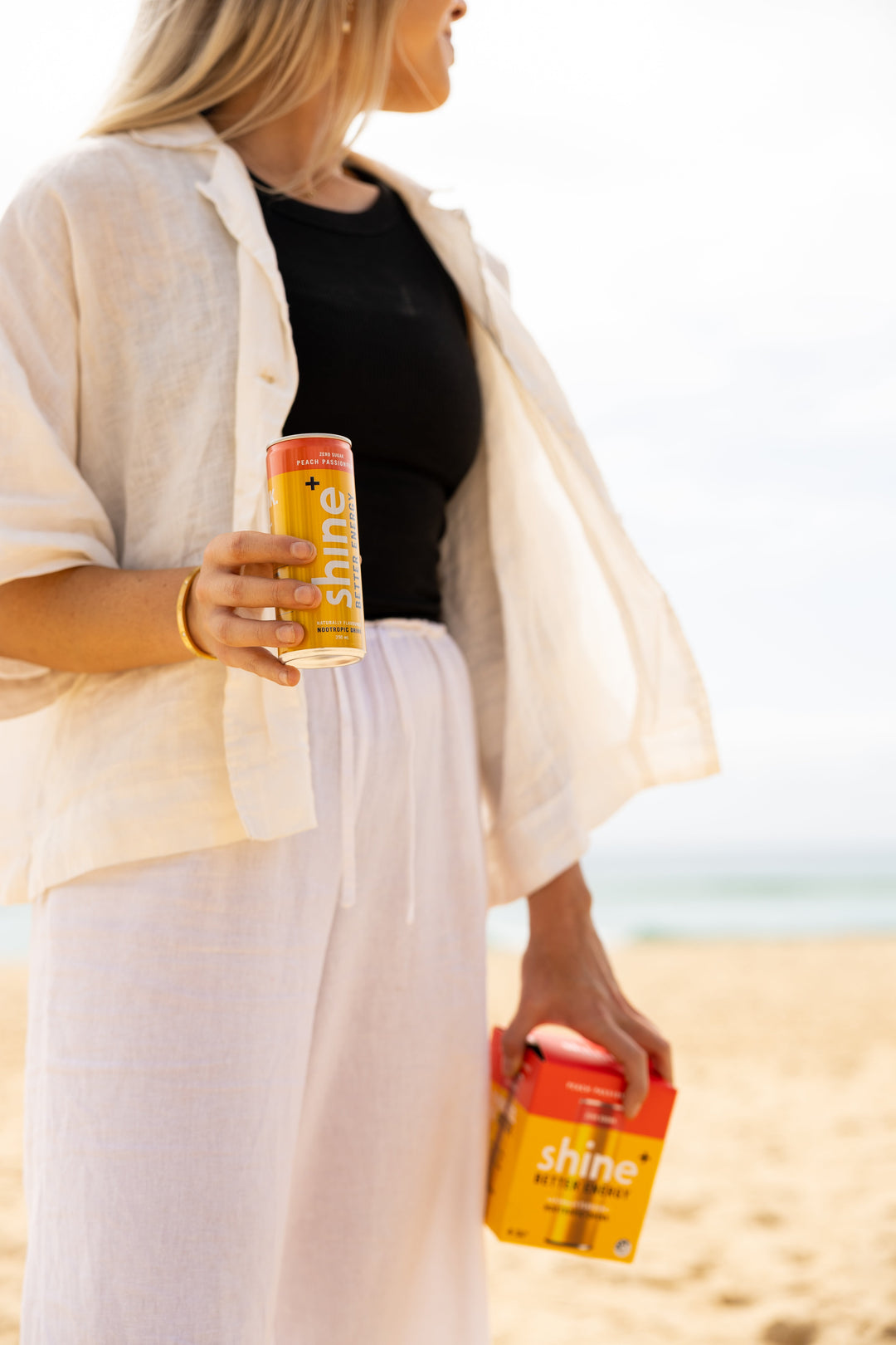Woman holding Shine+ peach passionfruit drink can and pack at the beach, bright sunny day