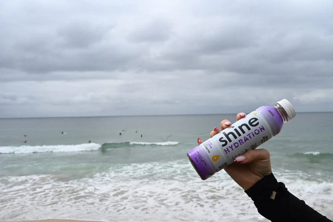 Hand holding grape Shine Hydration drink bottle by the ocean on a cloudy day