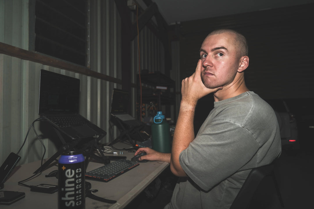 Person at desk with laptops, keyboard, and a Shine Charged drink can in a low-light office.