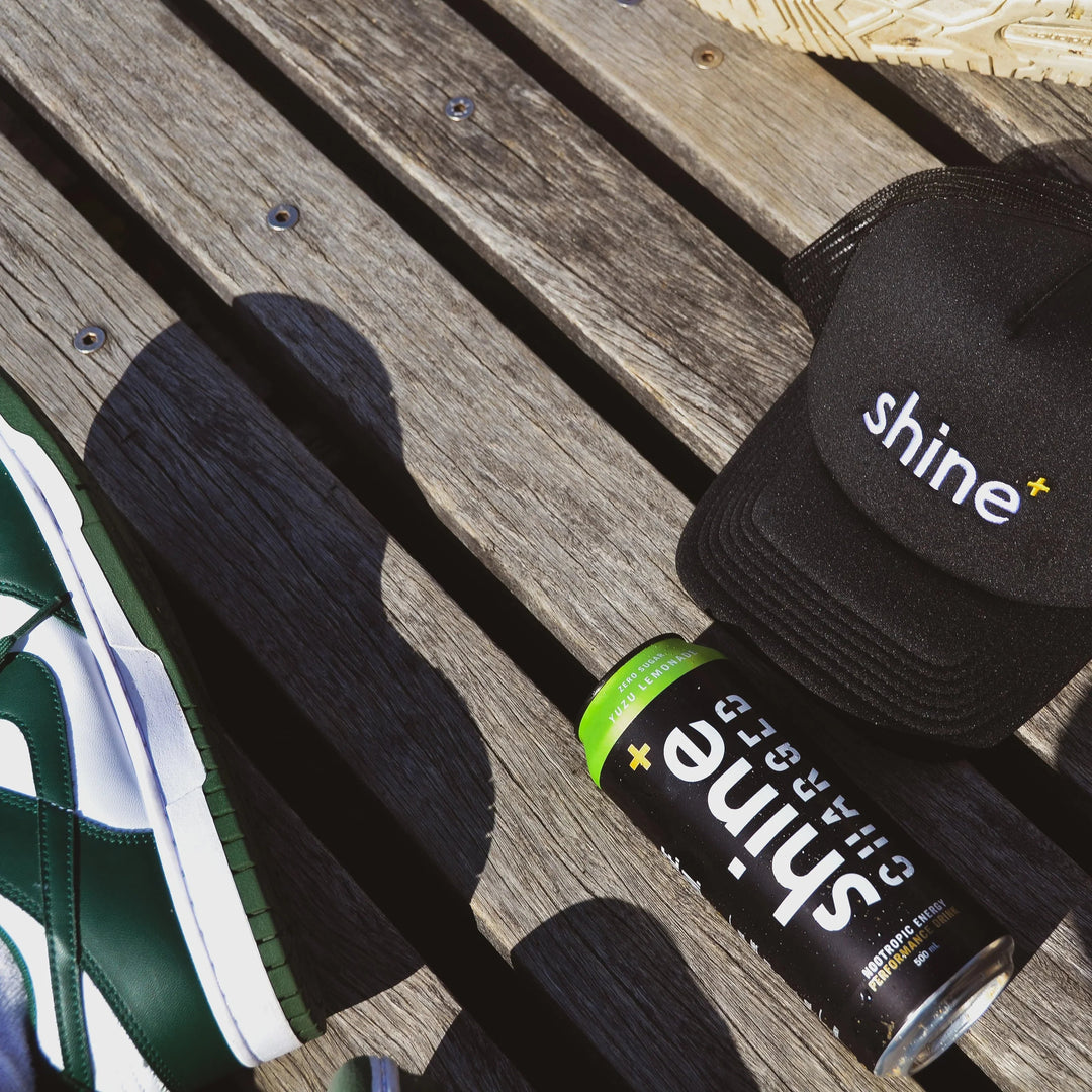 Shine+ nootropic energy drink can and branded cap on wooden bench, green sneakers visible