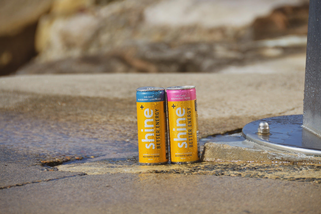 Two Shine nootropic energy drink cans, Blueberry Lemonade and Raspberry Soda, on wet pavement outdoors