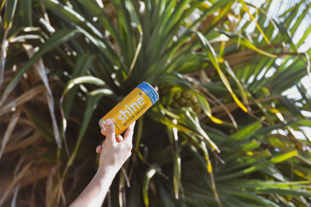 Hand holding yellow Shine Better Energy drink can outdoors with green tropical leaves