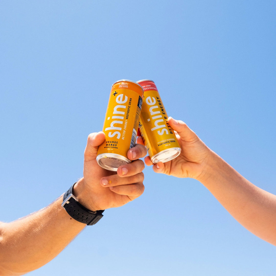 Two hands holding orange Shine nootropic drinks against a clear blue sky