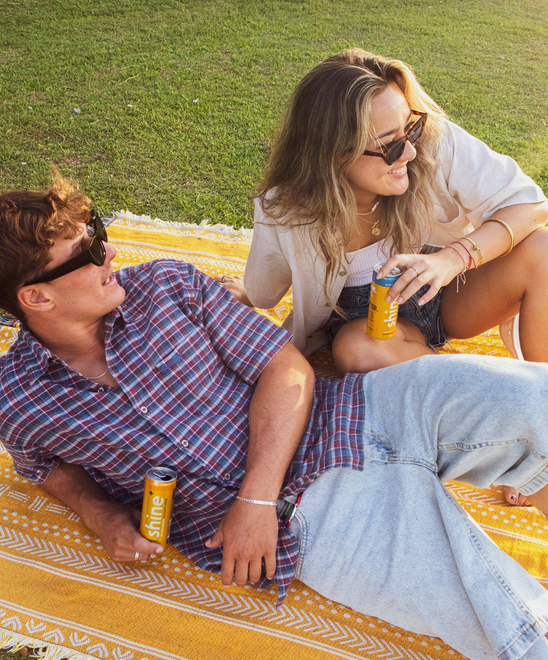Two people relaxing on a picnic blanket outdoors, holding yellow Shine+ drink cans, enjoying sunny weather
