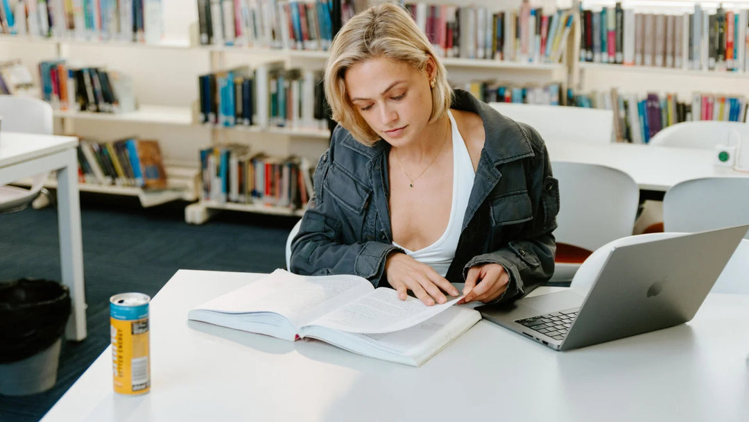 Woman studying with open book, laptop, and yellow energy drink can in a library setting