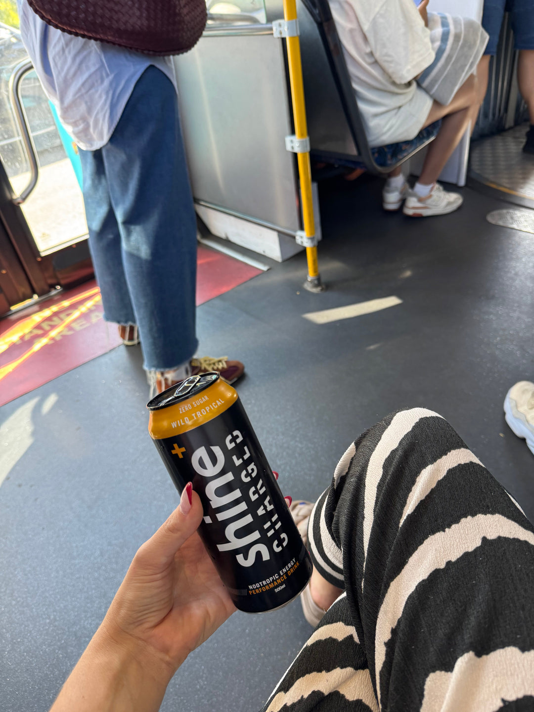 Hand holding Shine Charged nootropic energy drink can on a bus with people in casual wear.