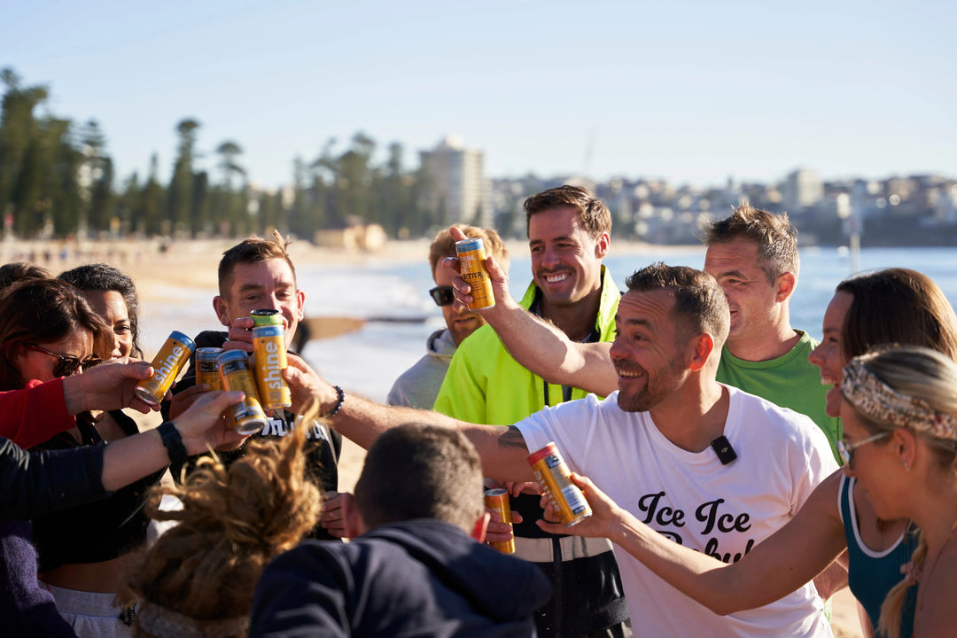 Group of people smiling and toasting Shine energy drink cans at the beach on a sunny day