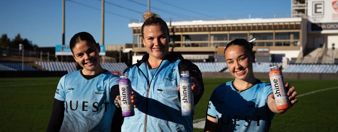 Three female athletes in blue jerseys holding Shine Hydration drinks on a stadium field