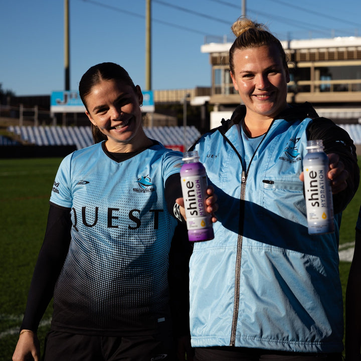 Two female athletes in blue sports uniforms holding Shine Hydration nootropic drinks outdoors